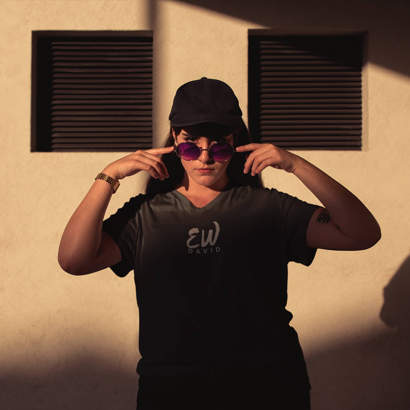 Woman wearing black baseball cap looks over her purple sunglasses while wearing an Ew David short sleeve t-shirt.