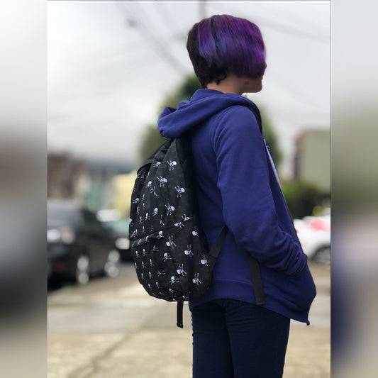 Tiny human with purple hair wearing skull and crossbones backpack facing away from the camera