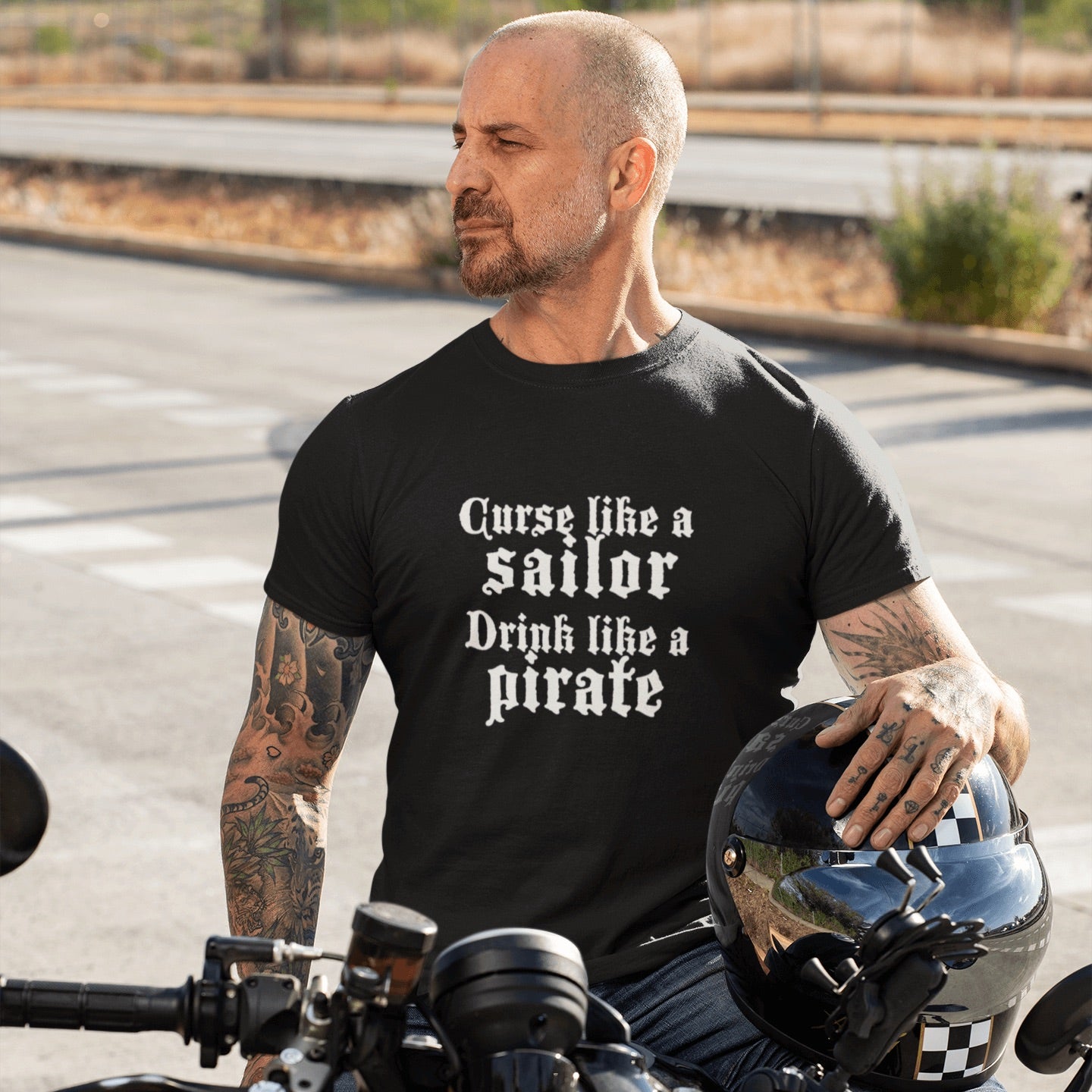Man with shaved head sits on motorcycle with hemet under his hand. He has tattoos and wears a black shirt with the words curse like a sailor drink like a pirate in white letters.