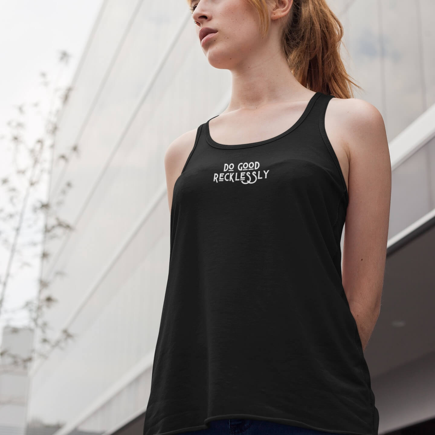 woman with strawberry blonde hair stands outside a building wearing a black do good recklessly relaxed racerback tank and looks off camera