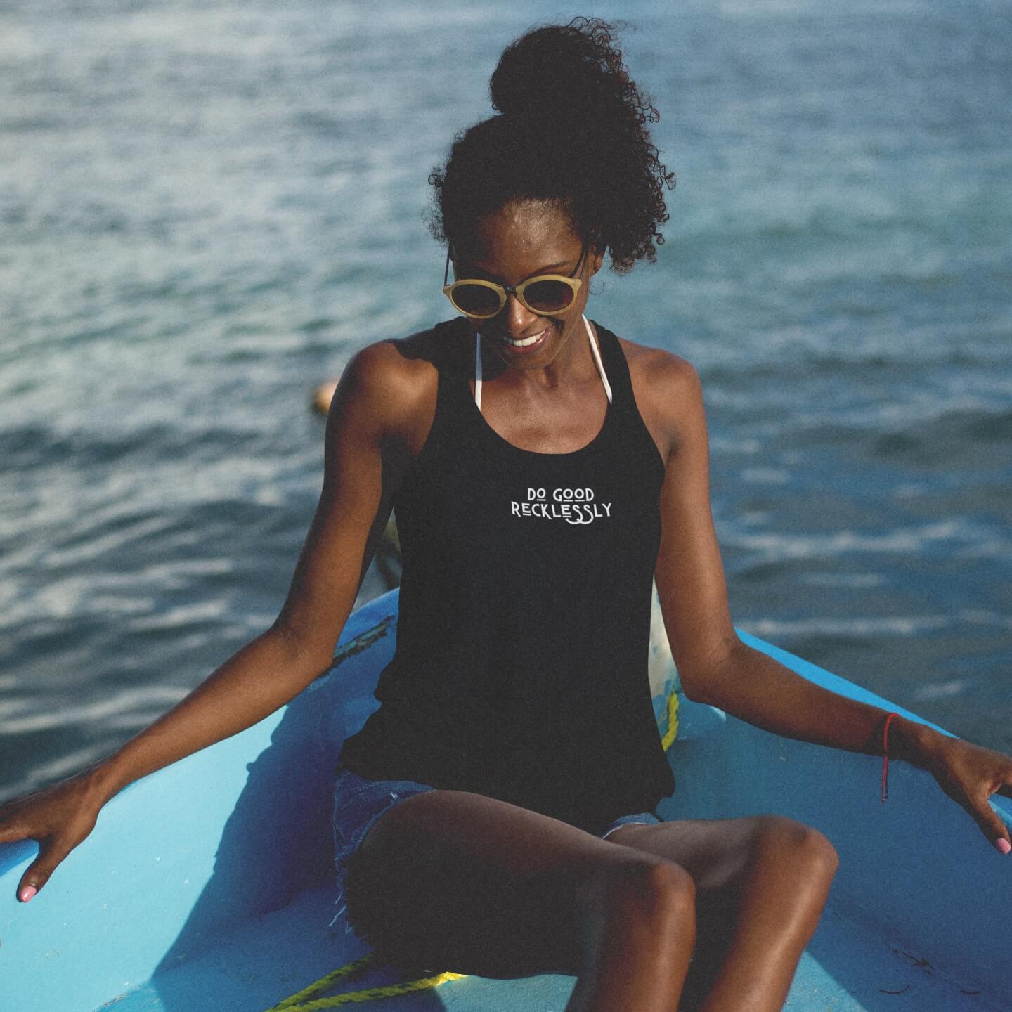 A Black woman with her hair in a high ponytail sits on a blue boat on the water wearing cutoff jean shorts and a black do good recklessly relaxed racerback tank.