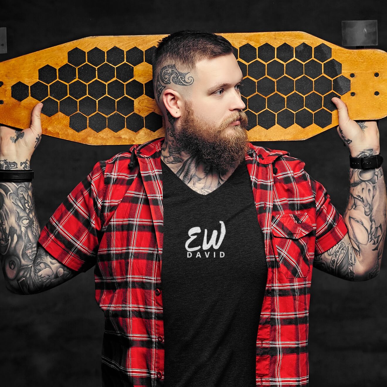 Man with tattoos holds a skateboard behind his head. He wears an Ew David short sleeve t-shirt with a red plaid flannel over it.