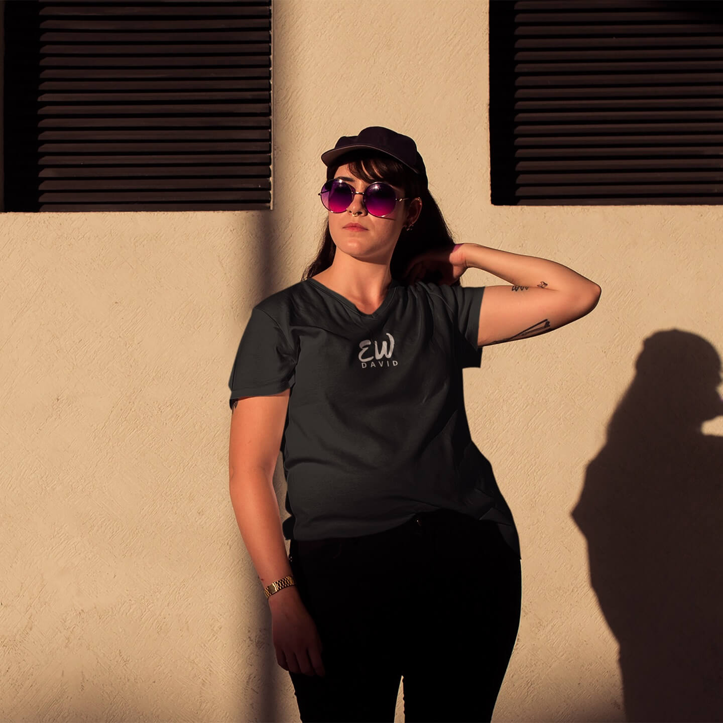 Woman in purple sunglasses and black baseball cap with black jeans and an Ew David short sleeve t-shirt in black looks into the distance.