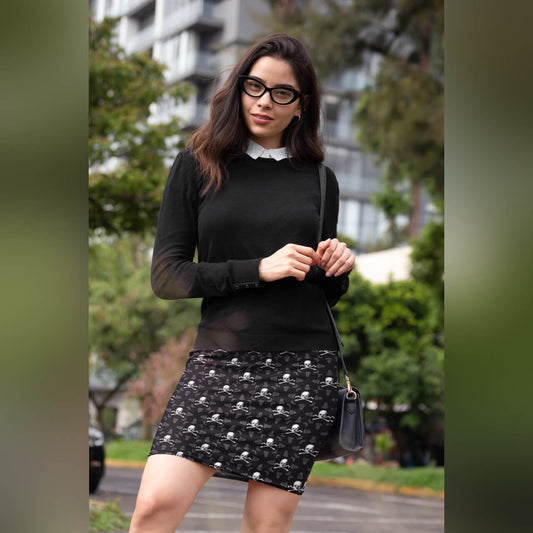 person with skull and crossbones pencil skirt, black sweater, and black rimmed glasses and long hair stand outside with purse.