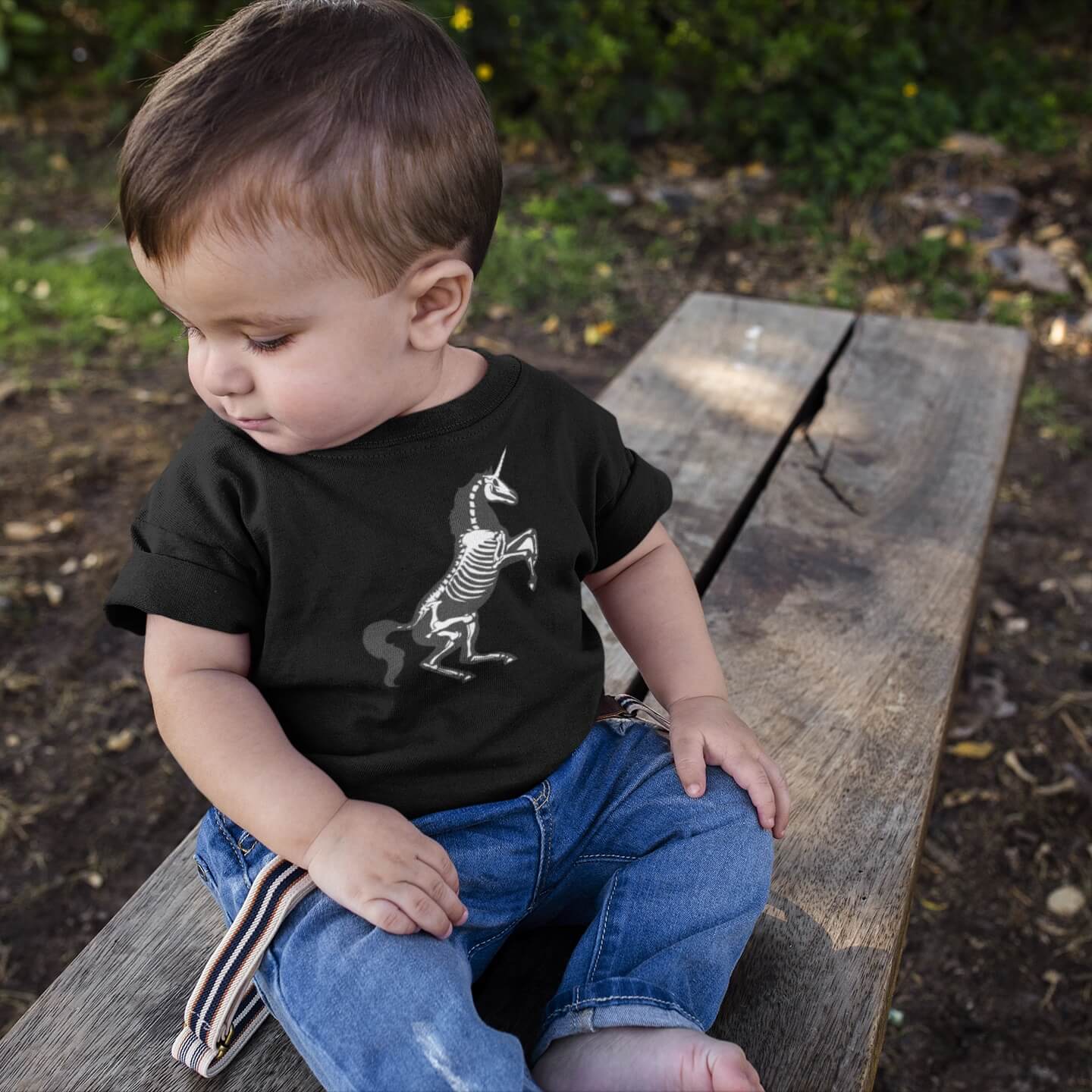 toddler with brown hair sits on a bench in jeans and a black unicorn skeleton baby onesie