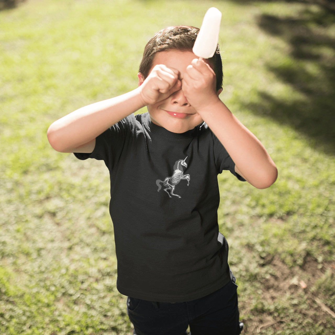 Brunette boy holds his fists in front of his eyes while also holding a white popsicle while standing on a grass lawn wearing a unicorn skeleton youth t-shirt in black